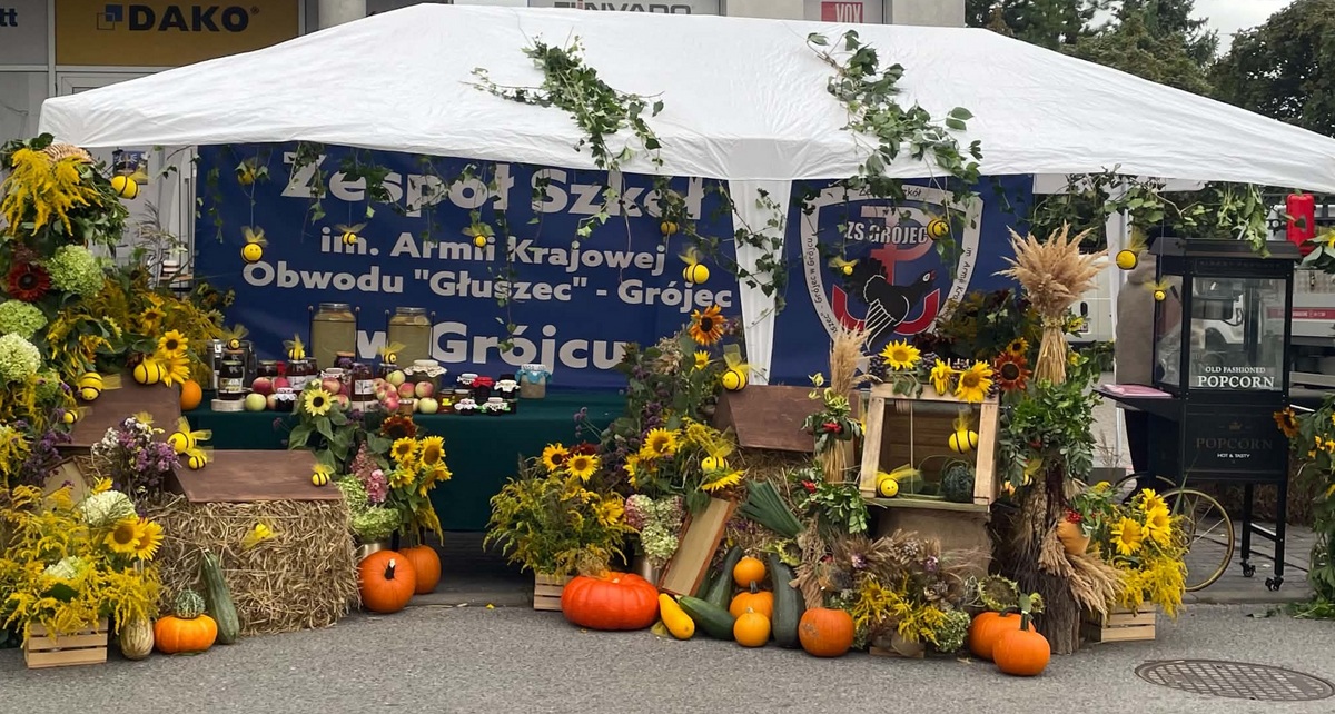 Stoisko Zespołu Szkół w Grójcu pod białym namiotem, udekorowane słonecznikami, dyniami, snopami zbóż, owocami i przetworami. W tle widoczny jest baner szkoły, a po prawej stoi wózek do popcornu.